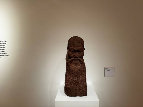 Скульптурный портрет старика с длинной бородой в тюбетейке из красновато-коричневого камня на белом постаменте у светлой стены музея. Справа висит табличка.