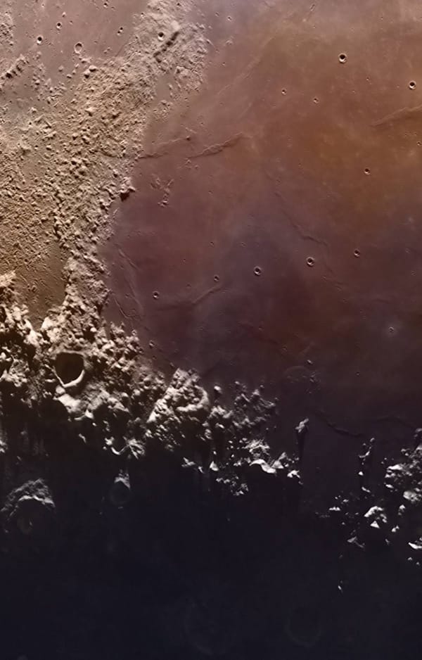 Крупный план лунной поверхности с кратерами, освещённой горной местностью внизу и тёмной областью в нижней части кадра, в тёплых оттенках.