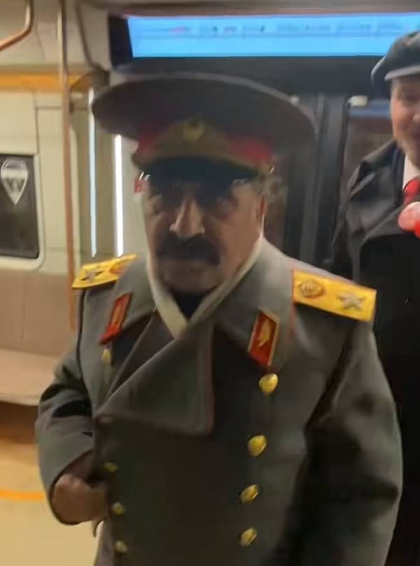 Усатый мужчина в сером мундире с золотыми погонами и в фуражке в вагоне метро, за ним человек в кепке и с красным галстуком.