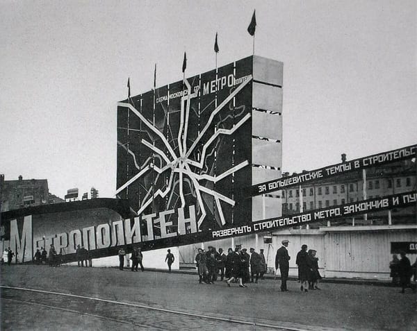 Огромный щит со схемой линий метро, надпись «Метрополитен» и ленты с лозунгами о темпах строительства. Перед ним идут прохожие, рядом дощатое ограждение стройки.