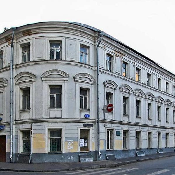 Старый оштукатуренный угловой дом с закруглённым фасадом, арочными наличниками окон и водосточными трубами. Рядом тротуар и знак «Въезд запрещён».