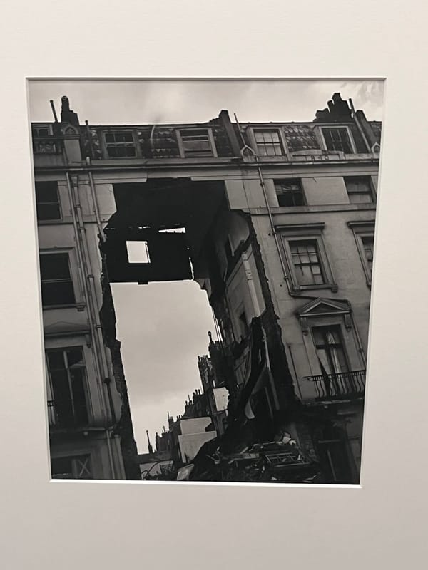 Чёрно-белая фотография на выставке: многоэтажный дом с выбитыми окнами, посреди фасада зияет провал до неба, внизу груда обломков.