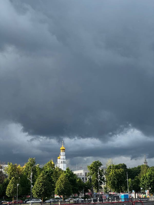 Тёмные грозовые тучи нависли над белой колокольней с золотым куполом. Внизу видны зелёные деревья, набережная с машинами и шпиль кремлёвской башни справа.