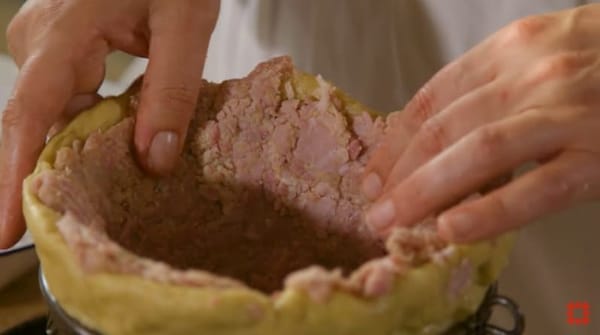 Крупным планом руки прижимают розоватый мясной фарш к стенкам формы из теста; внутри остаётся тёмная углублённая полость.