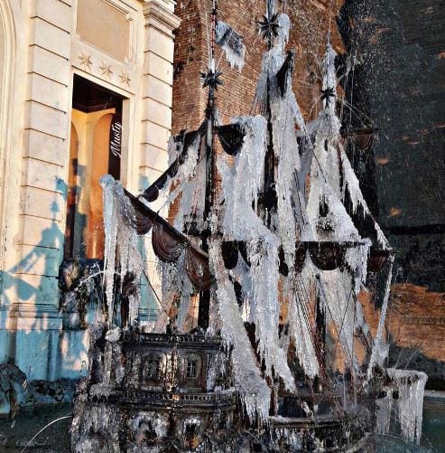 Фонтан в форме парусного корабля, мачты и снасти которого покрыты ледяной коркой и сосульками, на фоне здания и кирпичной стены.