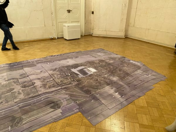 На паркетном полу старинного зала лежит наклеенная стопкой крупная аэрофотосъёмка с дорогой и зданием в центре. Слева идёт человек, у стены стоит белый прибор.