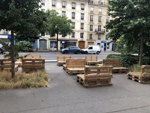 На тротуаре стоят скамьи и столы из деревянных поддонов среди деревьев и сухой травы. За ними улица с машинами и светлые многоэтажные дома.