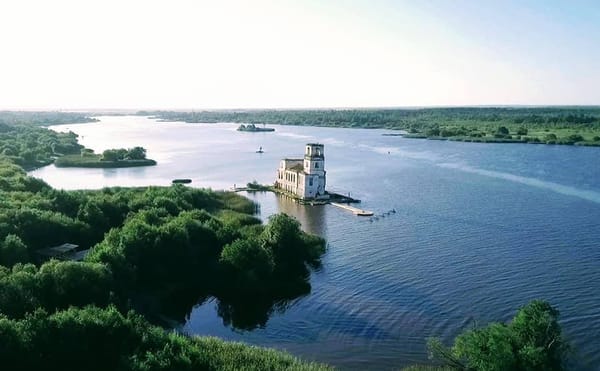 Аэросъёмка: белая церковь с колокольней стоит у воды на широком разливе реки. Вокруг зелёные острова и берега, вдали горизонт.