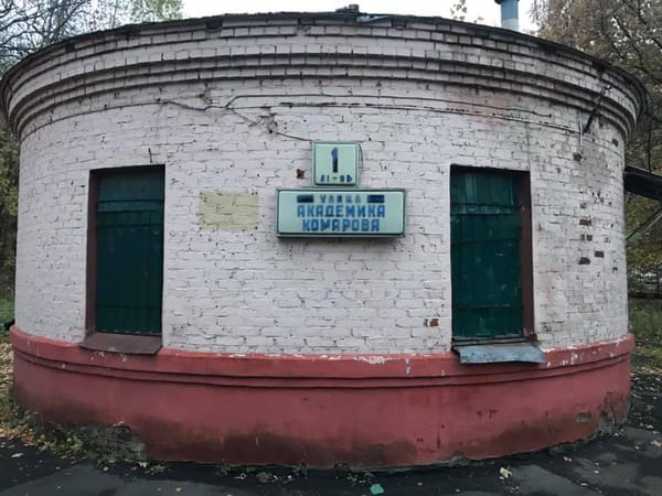 Круглое здание из побелённого кирпича с красным цоколем и двумя зелёными дверями. Между ними табличка: номер 1 и «Улица Академика Комарова». Вокруг осенняя листва.