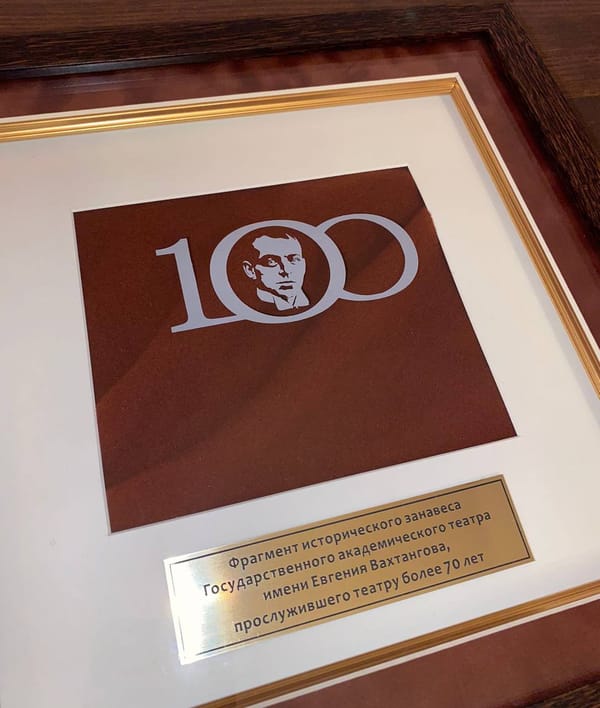 Рамка с фрагментом бордового занавеса, эмблемой «100» с портретом и золотистой табличкой о занавесе театра имени Вахтангова.