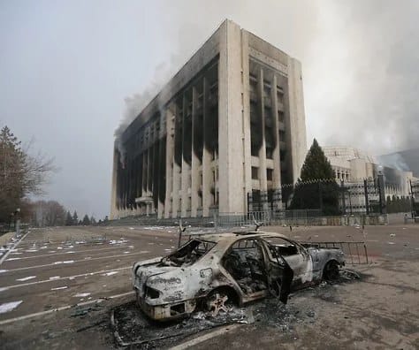 На пустой площади стоит обгоревший легковой автомобиль. За ним поднимается дым над многоэтажным зданием с закопчёнными окнами, рядом ограда и хвойное дерево.