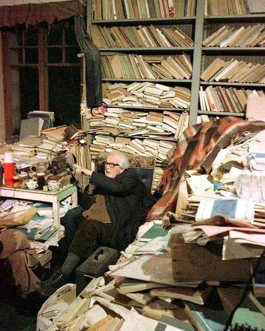 Пожилой мужчина в очках сидит в комнате среди стеллажей и груд книг и бумаг.