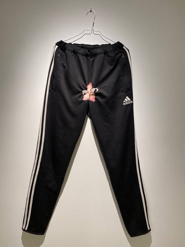 Чёрные спортивные брюки Adidas с белыми полосами висят на вешалке на стене, на уровне паха вышит розовый цветок.