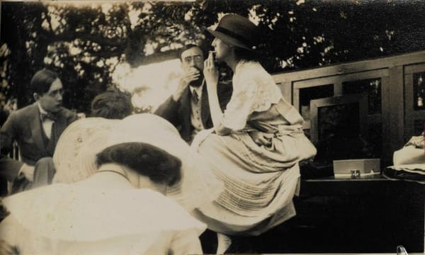 Старая сепиевая фотография: люди в одежде начала XX века сидят в саду, женщина в шляпе курит.