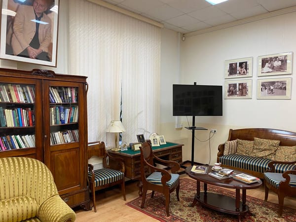 Комната с большим портретом мужчины на стене, застеклённым шкафом с книгами, письменным столом, диваном и креслами в полоску, журнальным столиком и чёрно-белыми фотографиями.