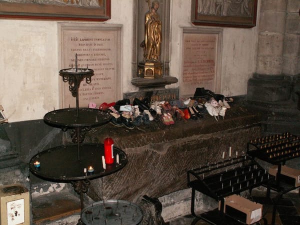 В соборе на каменной гробнице стоят ряды детской обуви, рядом подсвечники со свечами, а над ней золотая статуя святого и мемориальные таблички.