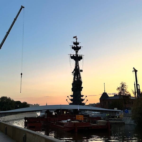 Силуэт памятника Петру I на фоне оранжево-голубого закатного неба. На переднем плане арочный мост, баржи с рабочими и стрела крана над водой.