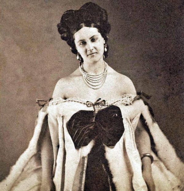 Старинная сепийная фотография: молодая женщина с высокой причёской, в серьгах и многорядном жемчужном ожерелье, в платье с открытыми плечами, меховой отделкой и большим тёмным бантом.